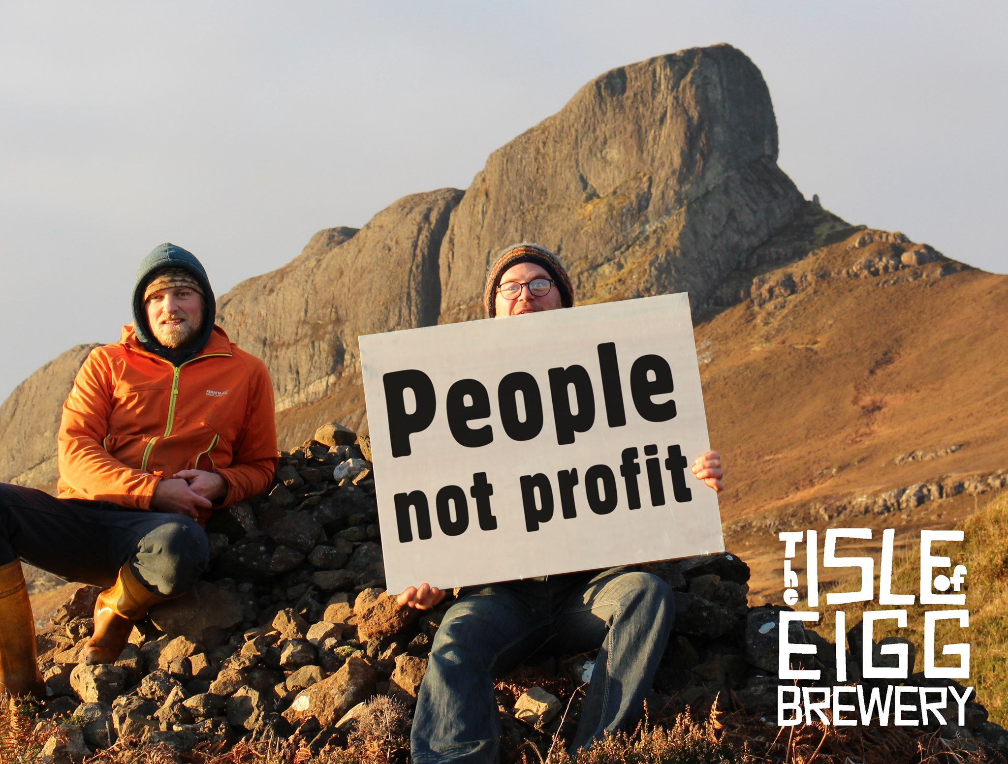 Founding members of Isle of Eigg Brewery pose before Sgurr, the island's highest peak, with a sign reading "People not Profit"